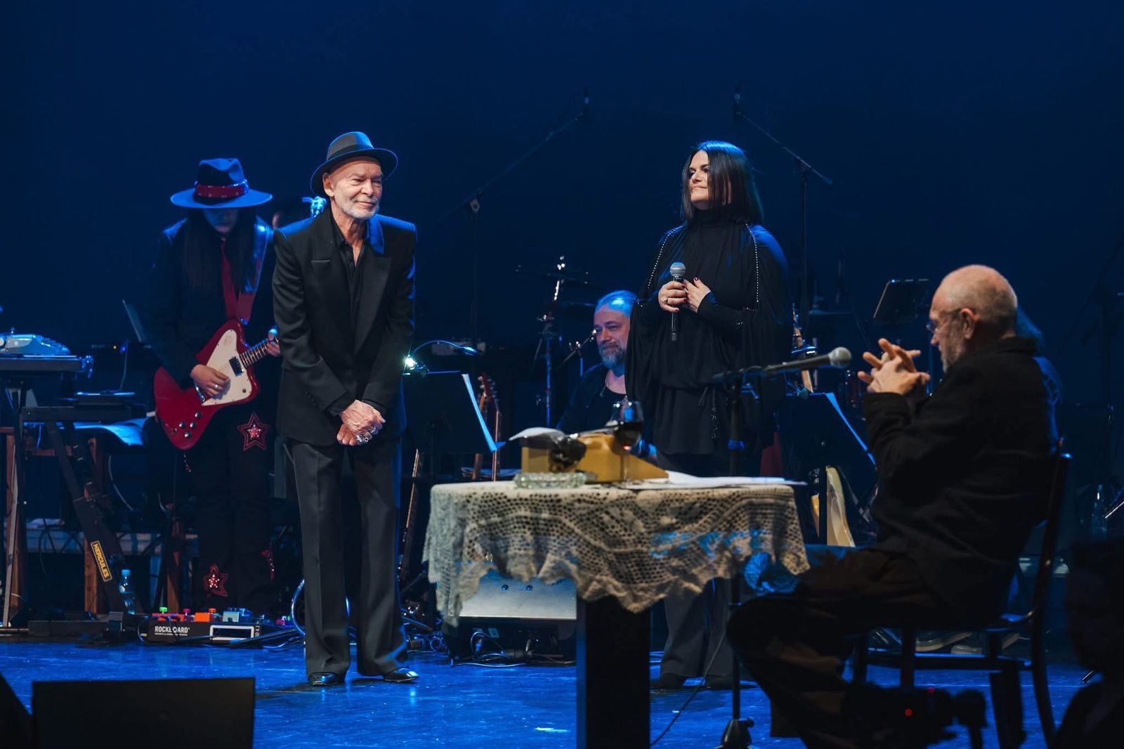 Sylwia Wiśniewska na scenie teatralnej z mikrofonem, obok wokalista i muzycy
